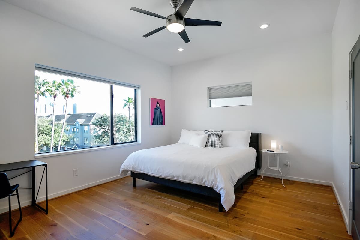 A bedroom with a bed, a desk, a chair, a ceiling fan, a window with a view of houses and trees, and a bedside table with a lamp.