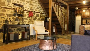 Un intérieur rustique avec un mur en pierre, un escalier en bois, une chaise blanche, une table basse avec un plateau en verre et un coin cuisine.