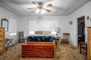 A bedroom with a bed, a wooden chest, a chair, a desk, a lamp, and a ceiling fan.