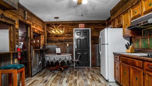 Une cuisine rustique aux murs en bois, avec un réfrigérateur blanc et une table à manger recouverte d’une nappe en dentelle.