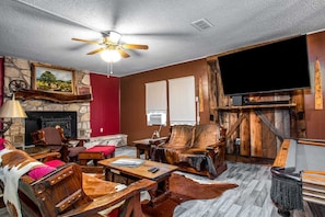 A rustic living room with a stone fireplace, wooden furniture, a ceiling fan, and a large flat-screen TV.