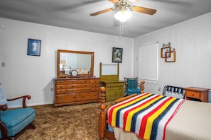 A bedroom with a wooden dresser, a bed with a striped blanket, a chair, a desk, and a ceiling fan. - 3H Ranch Mountain Retreat | 60-Acre Ranch (Medina)