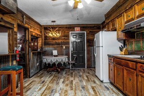 Une cuisine rustique aux murs en bois, avec un réfrigérateur blanc, une table en bois recouverte d’une nappe en dentelle et un lustre.