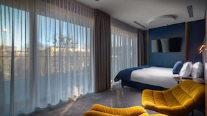 A modern hotel room with a large bed, a flat-screen TV, a yellow armchair, and a view of the city through sheer curtains. - My Story Gdynia (Gdynia)