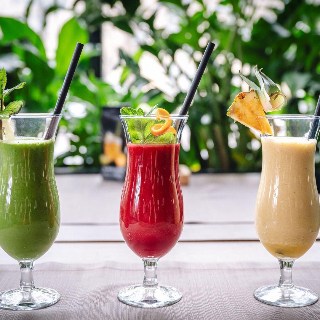 Trois verres contenant des boissons, chacun muni d’une paille et décoré de feuilles de menthe et de fruits, sont posés sur une table en bois. L’arrière-plan est flou et de couleur verte.