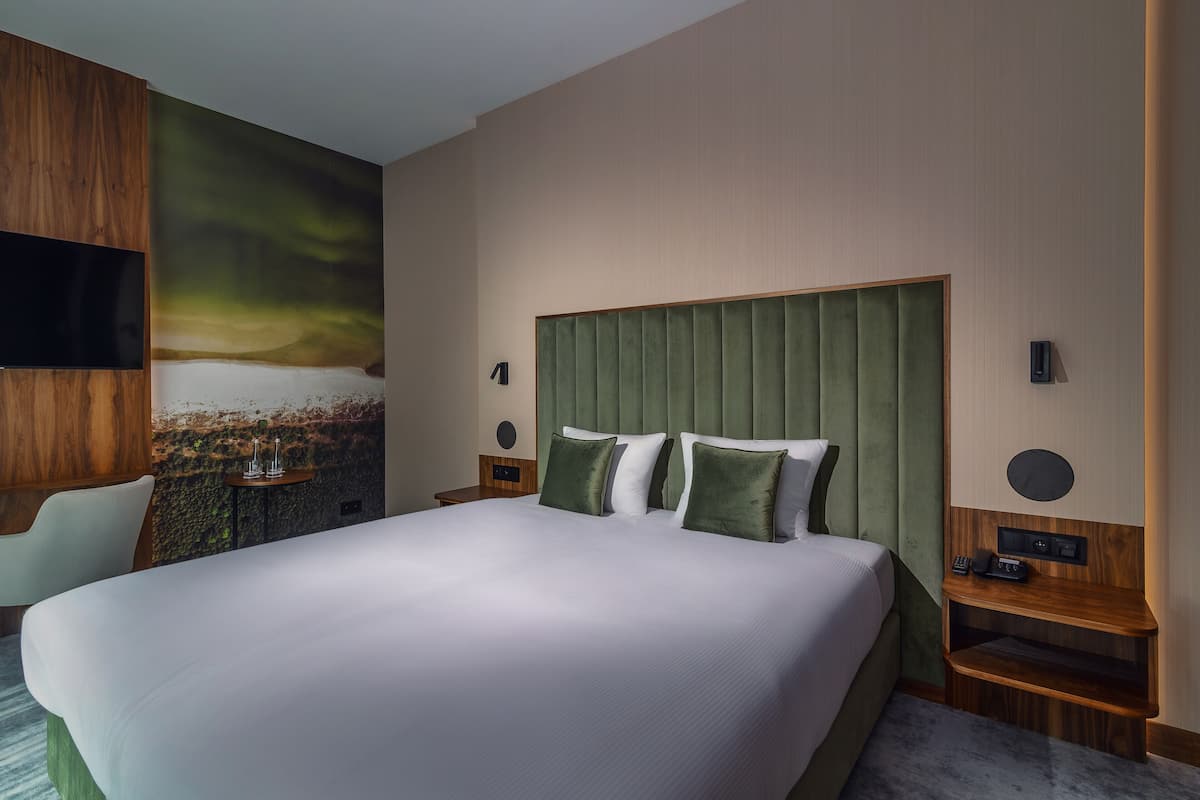 A modern hotel room with a large bed, green headboard, and a mural on the wall.