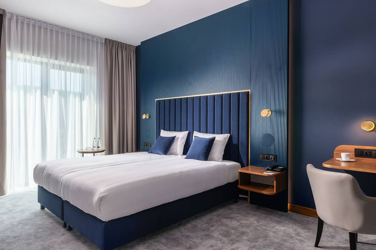 A modern hotel room with a large bed, a bedside table, a chair, and a desk with a cup.