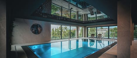 A modern indoor swimming pool with a glass ceiling, large windows, and a view of the outdoors.