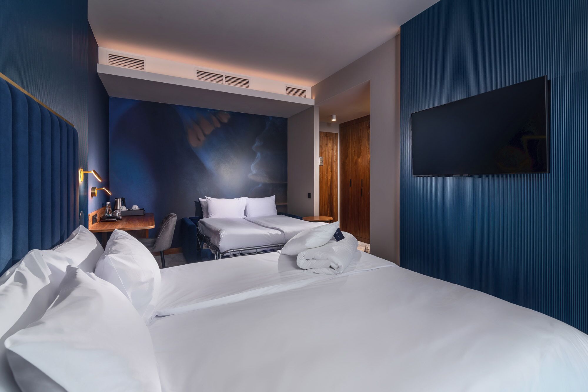 A hotel room with two beds, a desk with a lamp, a TV, and a large window.