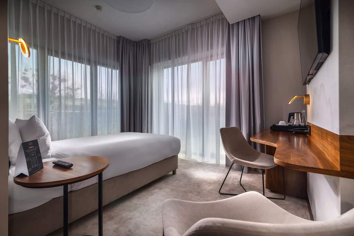 A modern hotel room with a large bed, a desk with a chair, a small table, and a TV mounted on the wall.