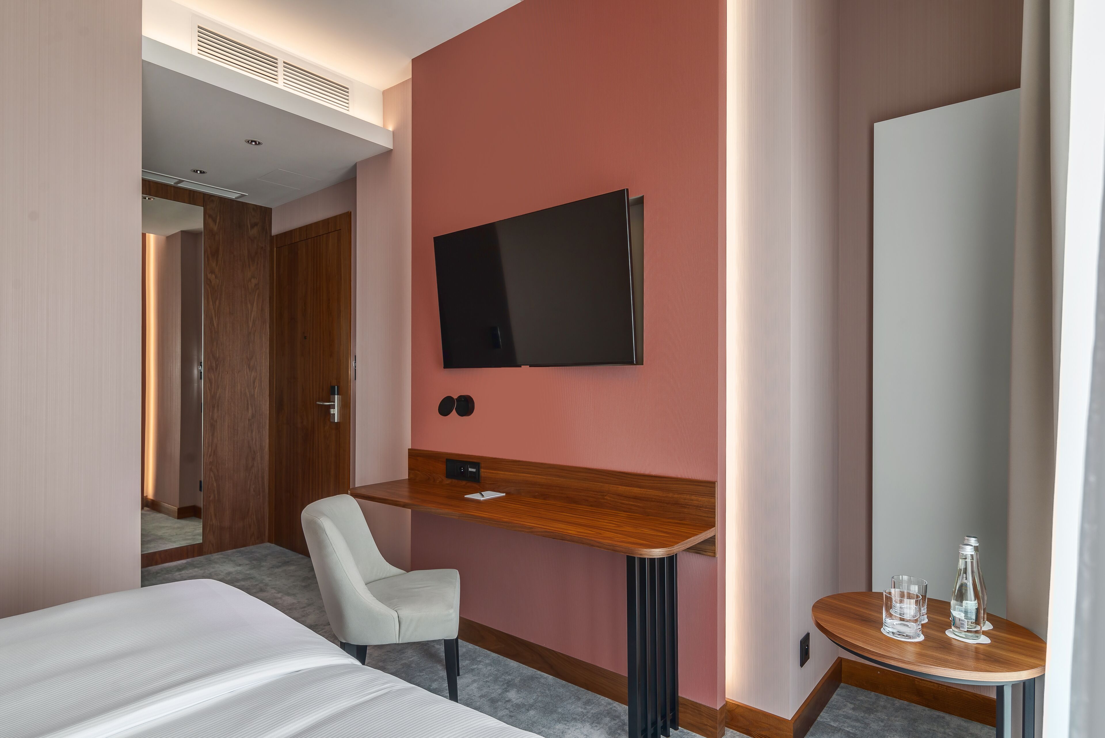 A hotel room with a bed, a desk, a chair, a TV, and a small table with glasses and a bottle.