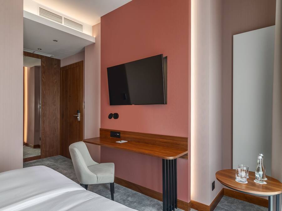 Habitación de hotel con cama, escritorio, silla, televisión y una pequeña mesa con vasos y una botella.