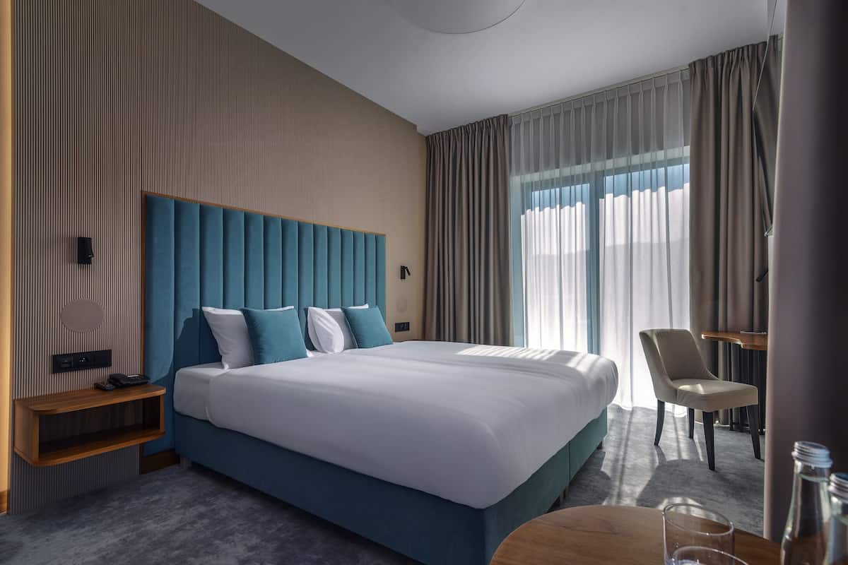 A modern hotel room with a large bed, a bedside table, a chair, and a small table with a glass and bottle.