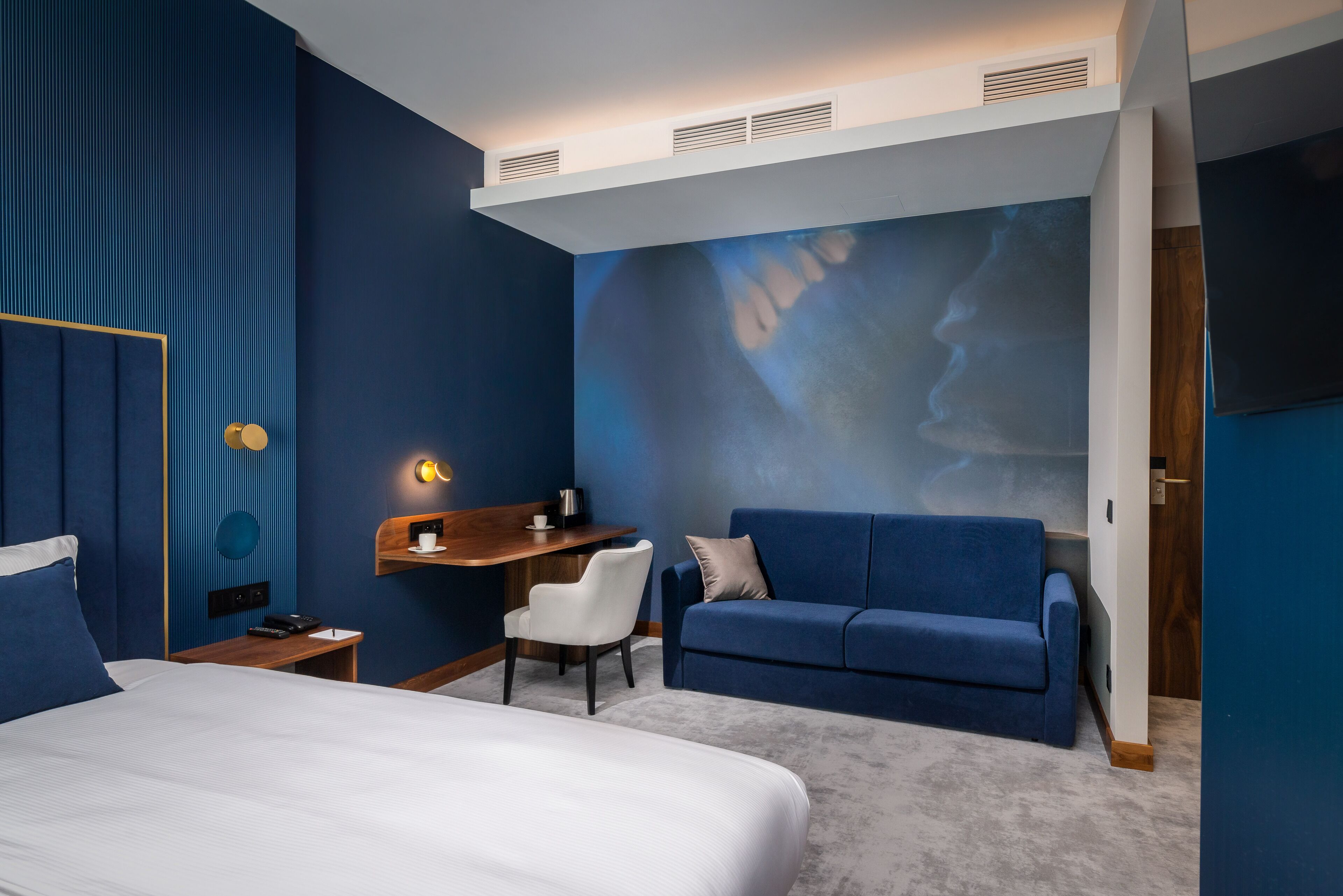 Quarto de hotel moderno com uma cama grande, um sofá azul, uma escrivaninha com cadeira e uma tela projetora fixada na parede.