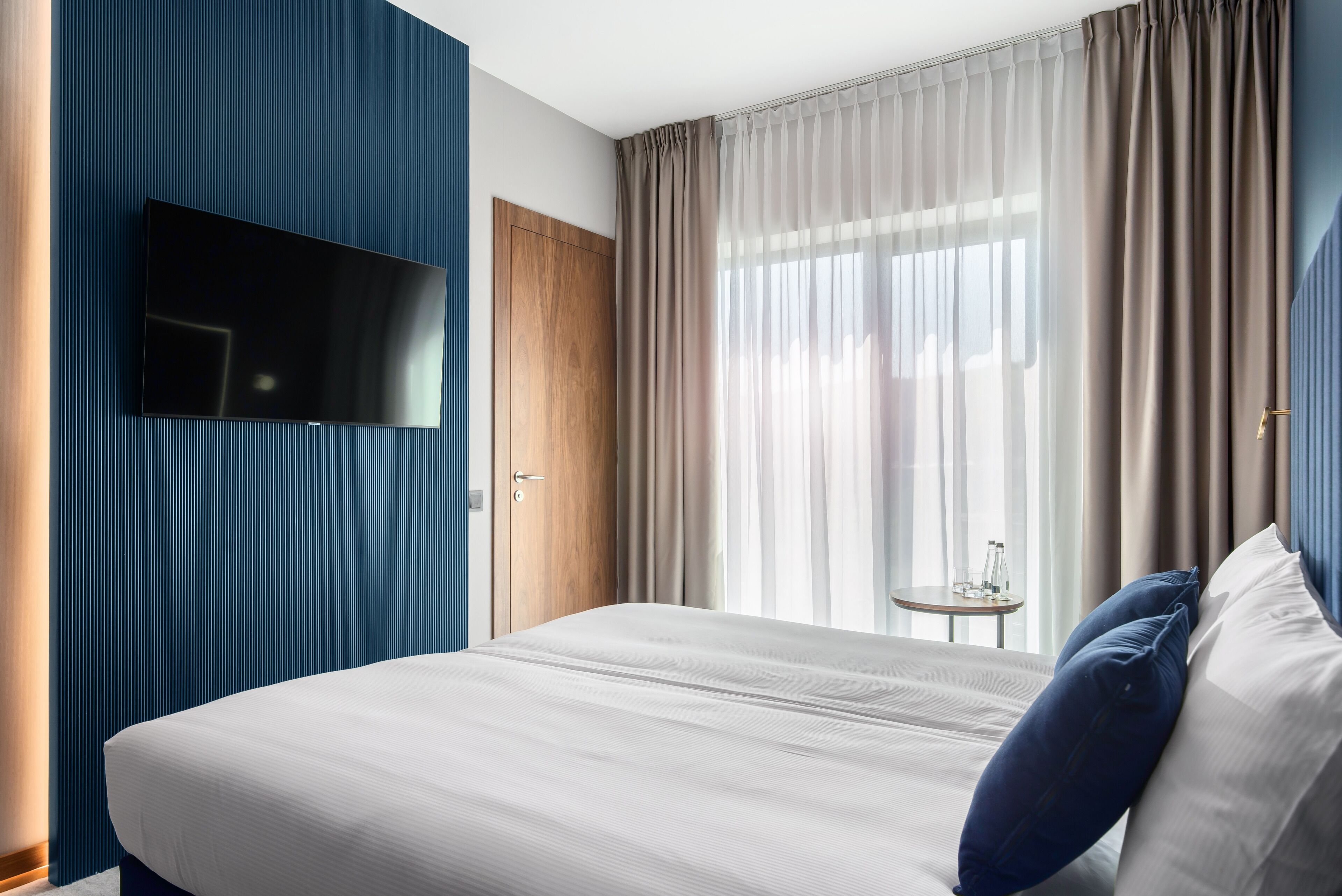 A modern hotel room with a large bed, a flat-screen TV mounted on the wall, a wooden door, and a small table with a lamp.