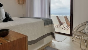A bedroom with a large bed, a view of the beach, a wicker chair, and a chandelier.
