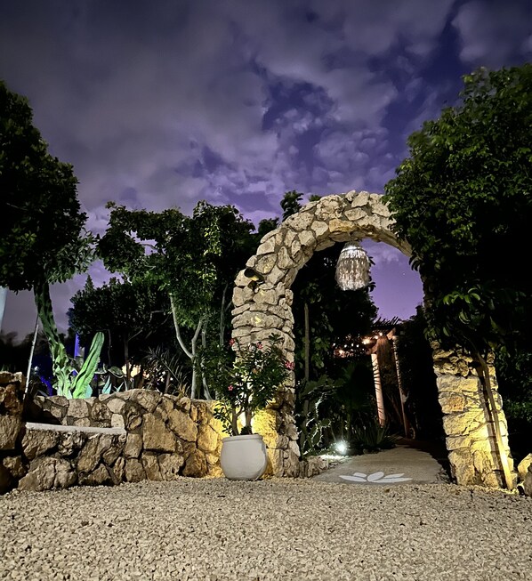 An outdoor stone archway with a pathway leading through it, surrounded by greenery and a decorative hanging light fixture.