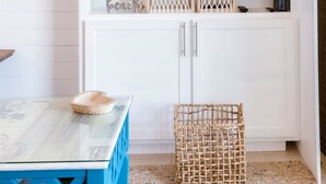 A modern kitchen with a large flat-screen TV mounted above a white cabinet, a blue table with a woven basket, and a patterned rug. - Stunning Ocean Views STEPS from the Beach with Pool! Fabulous 4th Floor Studio! (New Smyrna Beach)