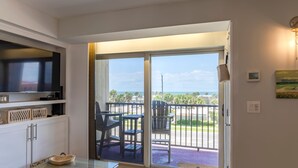 A room with a TV, a small table, and a chair, a sliding glass door leading to an outdoor area with a view of the ocean and palm trees. - Stunning Ocean Views STEPS from the Beach with Pool! Fabulous 4th Floor Studio! (New Smyrna Beach)