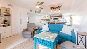A bright living room with a blue sofa, a white armchair, and a glass coffee table. There is a kitchen in the background with a dining table and chairs. - Stunning Ocean Views STEPS from the Beach with Pool! Fabulous 4th Floor Studio! (New Smyrna Beach)