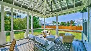 A covered outdoor deck with seating, a ceiling fan, and a view of a green lawn and house in the background. - 4 Bedroom Gorgeous Beachy Home - Resort Pool - 6 Seater Golf Cart (Santa Rosa Beach)