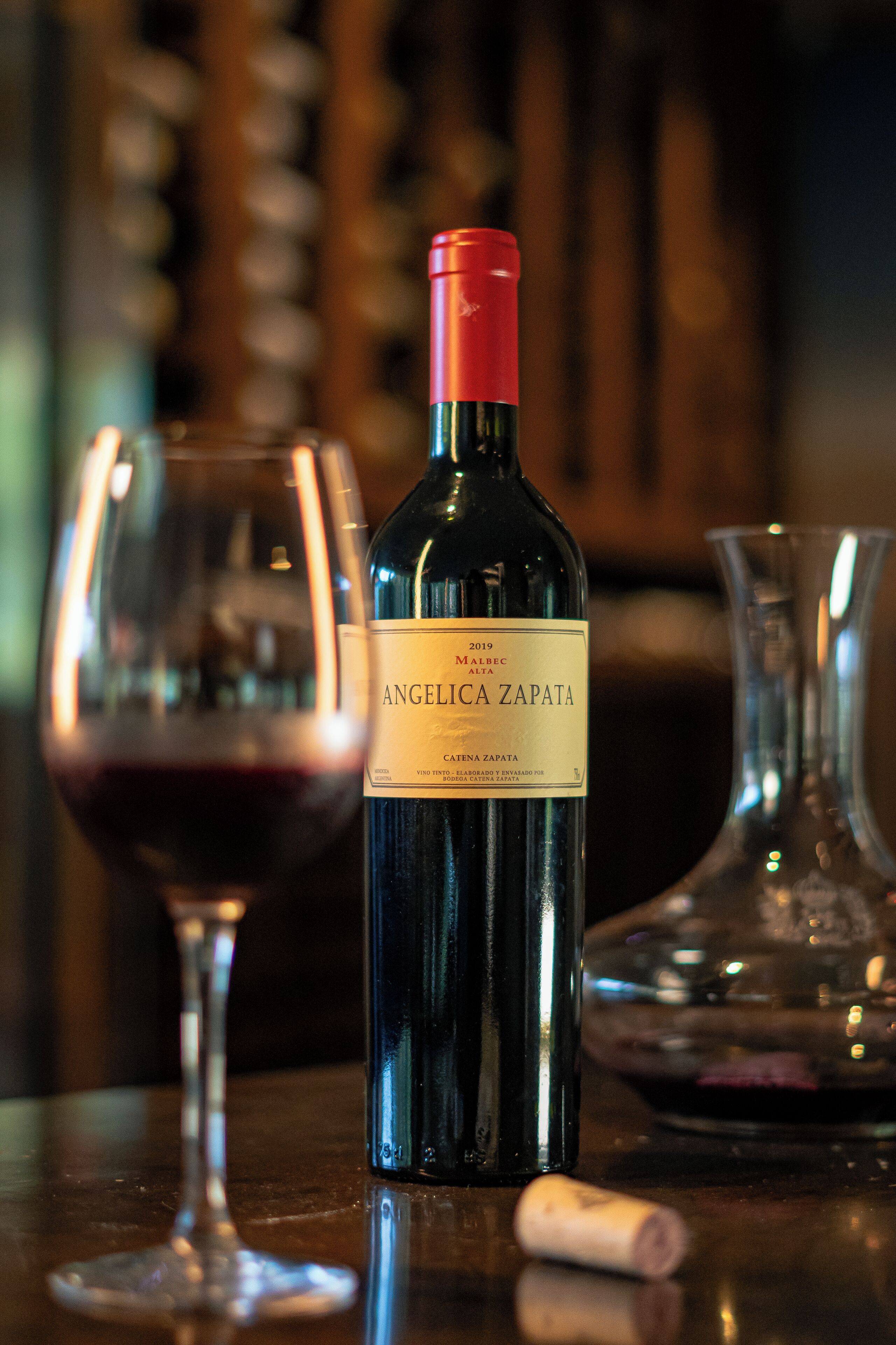 A bottle of Angelica Zapata wine, a glass of red wine, and a glass decanter on a wooden table.