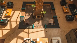 Un hall d’entrée spacieux avec une grande cheminée en briques au centre, plusieurs coins salon et des plantes en pot.