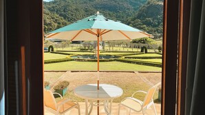 Une terrasse avec une table et des chaises, un parasol, et une vue sur un jardin aménagé avec une montagne en arrière-plan.