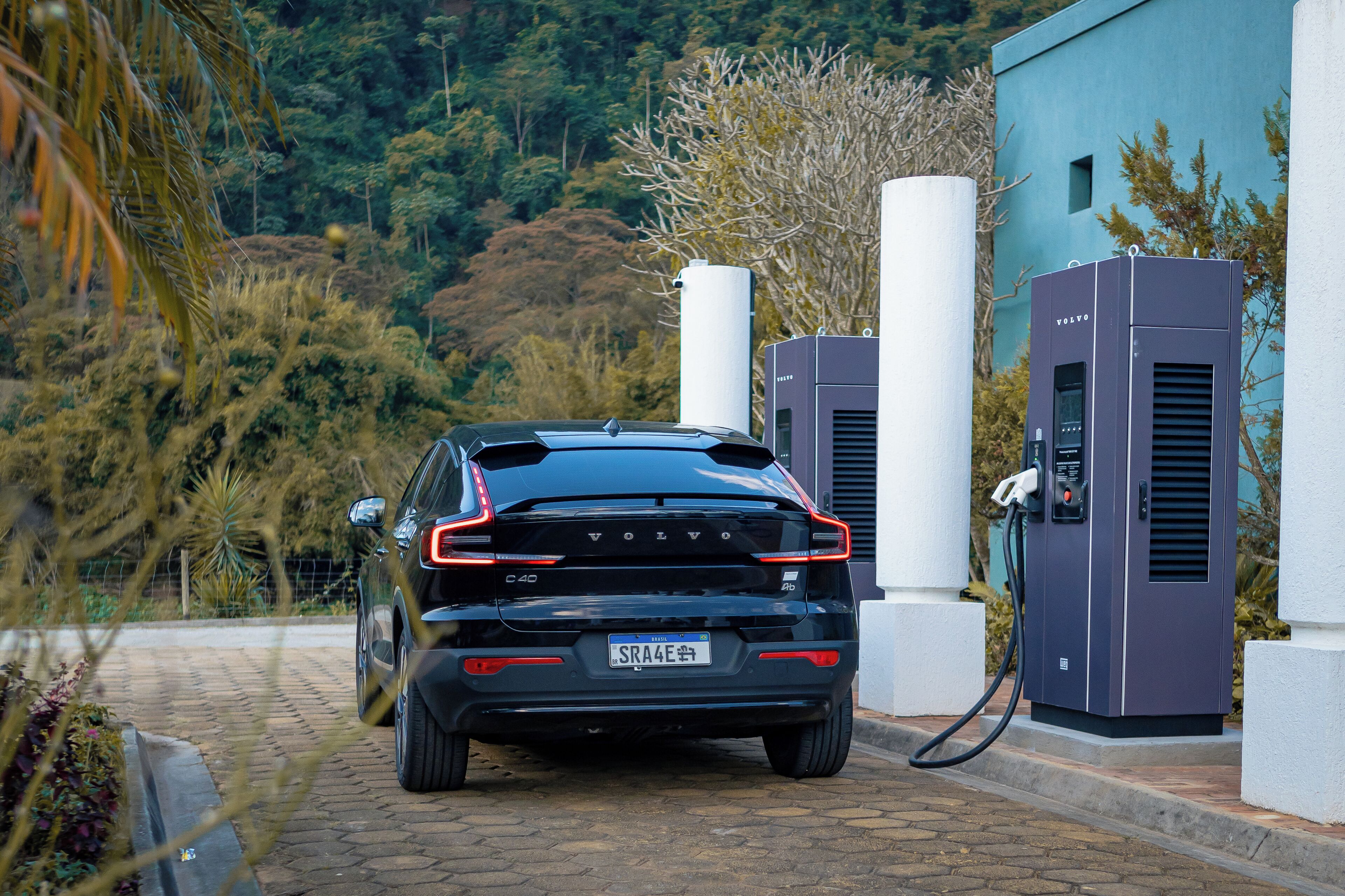 Un coche Volvo C40 negro estacionado en una estación de carga con dos cargadores eléctricos de gran tamaño, en un área ajardinada con árboles y un edificio al fondo.