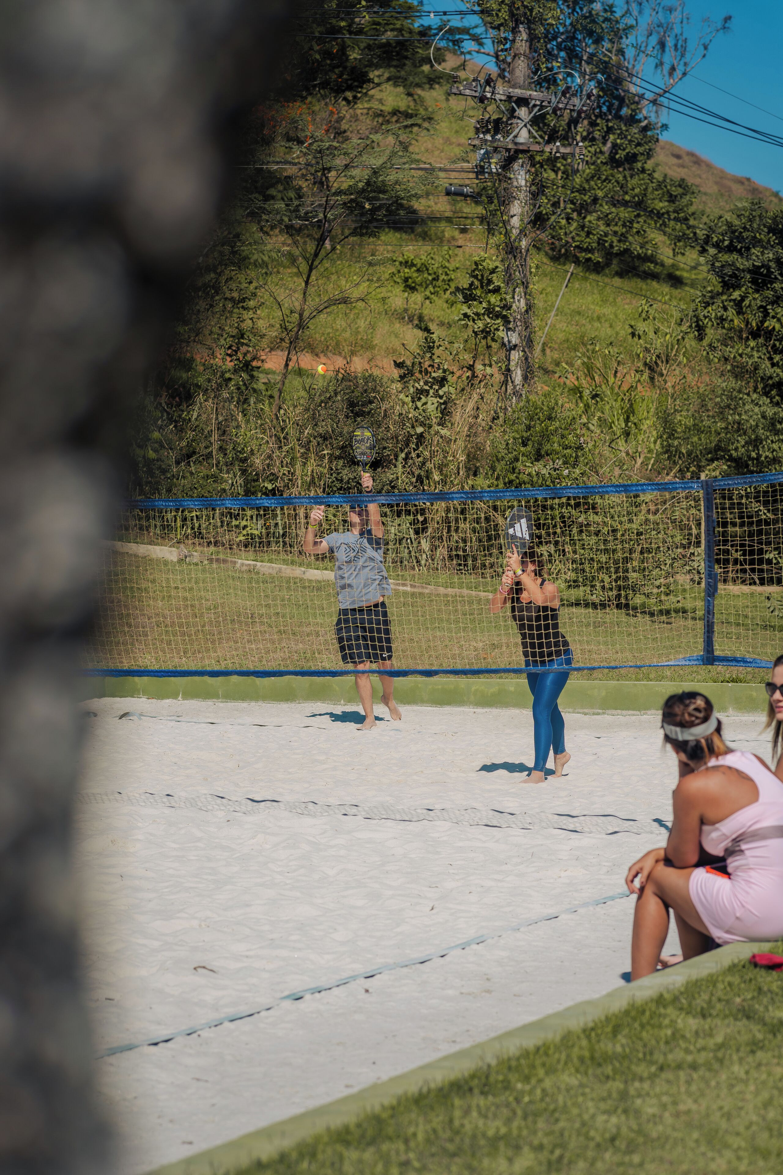 A volleyball game on a sandy court with a blue fence and greenery in the background.