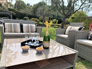Eine Terrasse mit Korbmöbeln, einem Glastisch und einer Champagnerflasche.