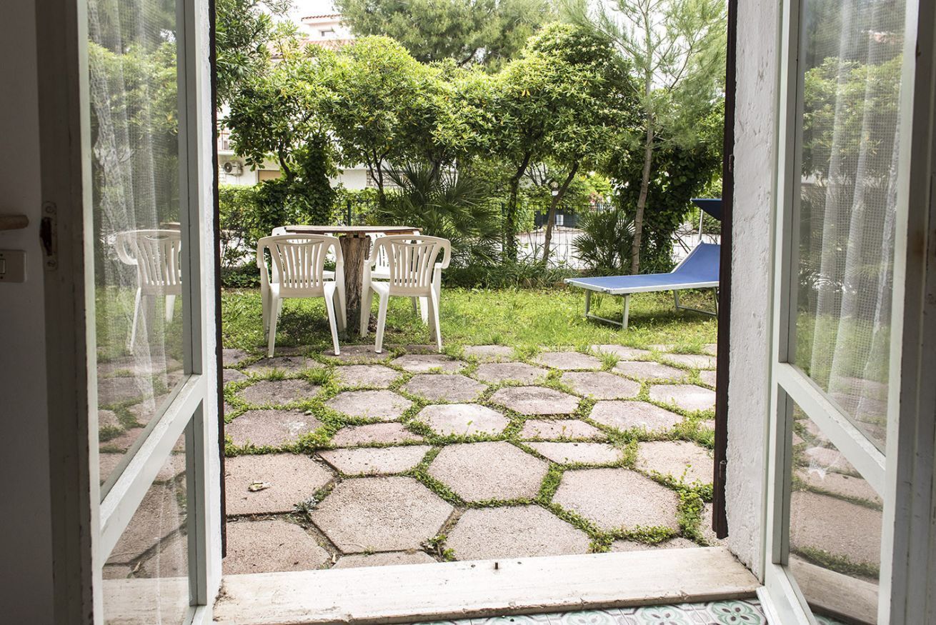 Porta aperta che dà su un patio con piastrelle di pietra esagonali, due sedie di plastica bianca e un tavolo blu.