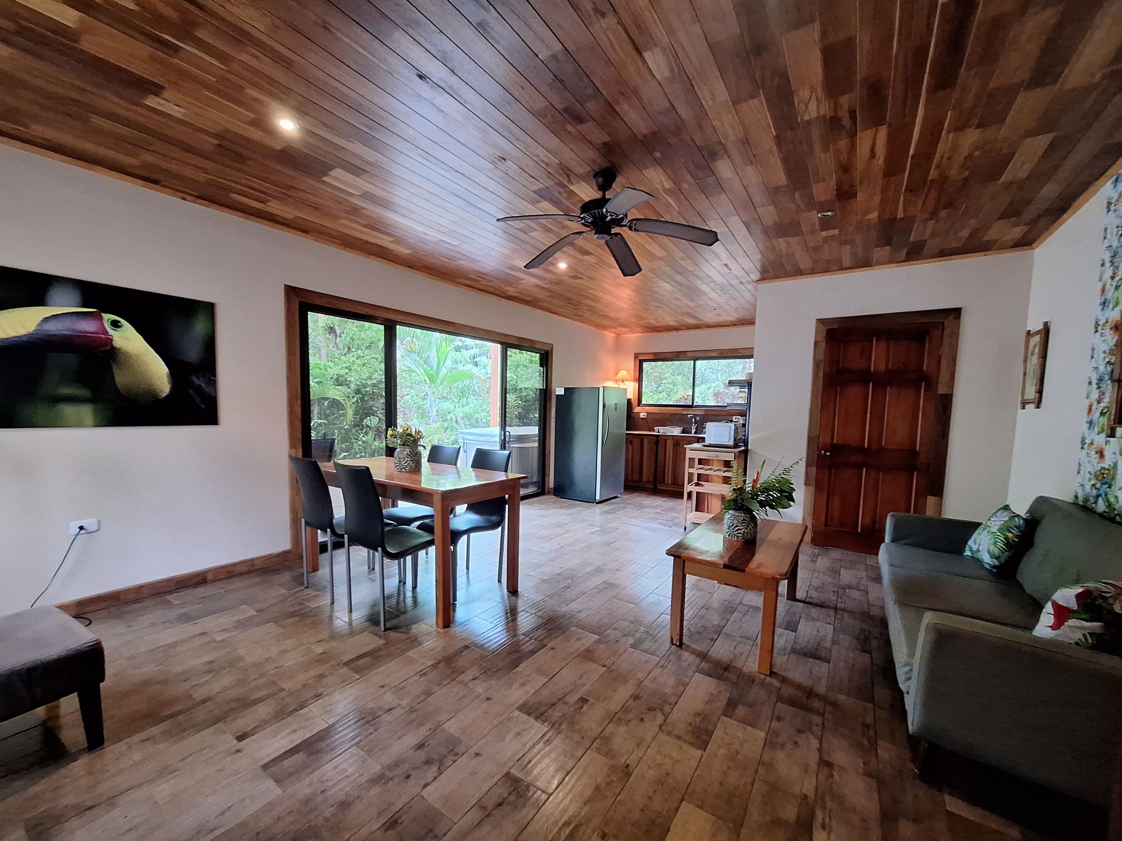 A spacious living area with a wooden ceiling, a dining table with chairs, a sofa, and a kitchen area visible through a glass door.