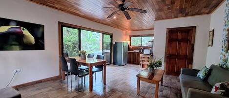 A spacious living area with a wooden ceiling, a dining table with chairs, a sofa, and a kitchen area visible through a glass door.