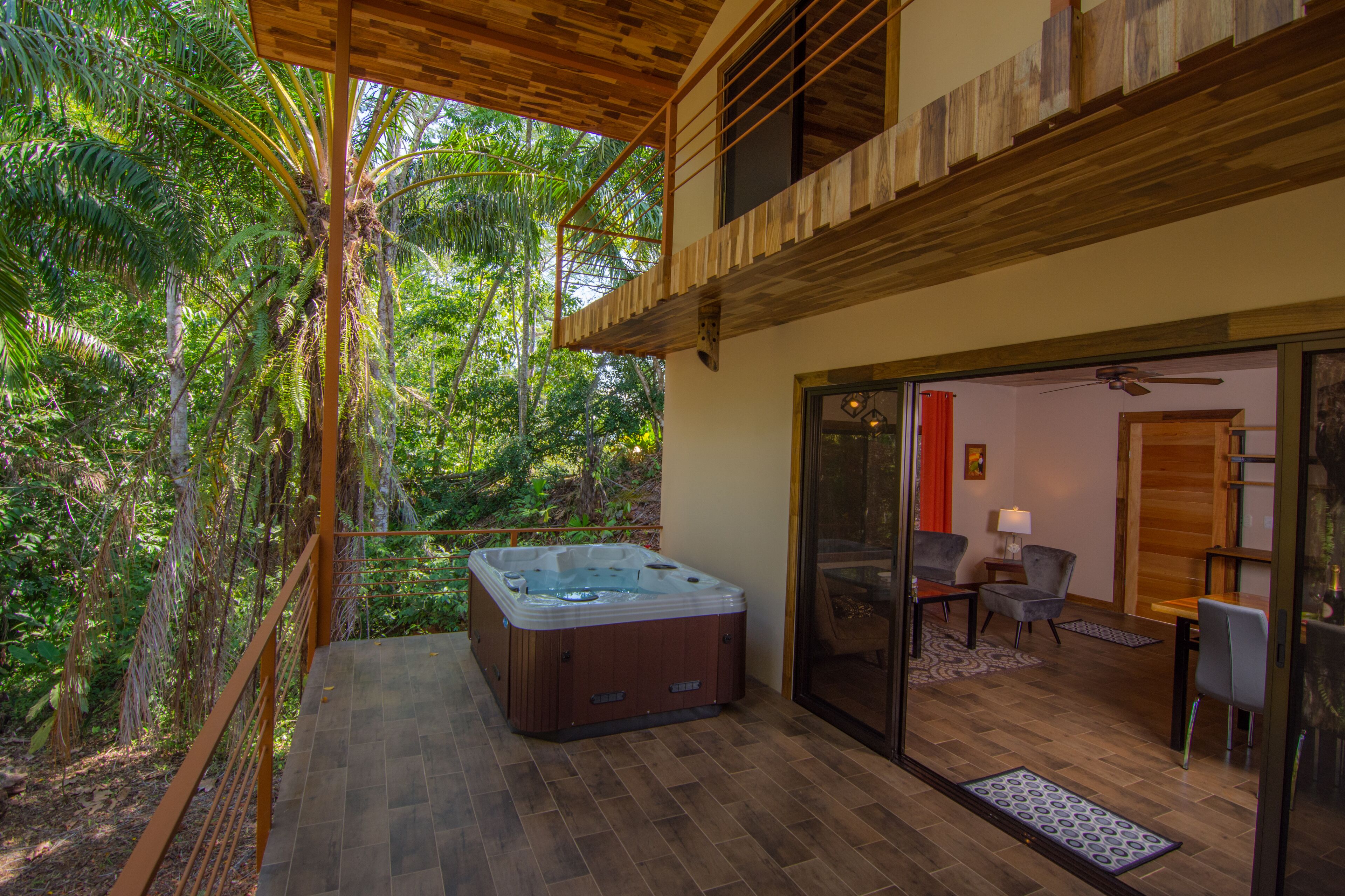 A hot tub on a wooden deck with a glass door leading to a room with a chair and a table.