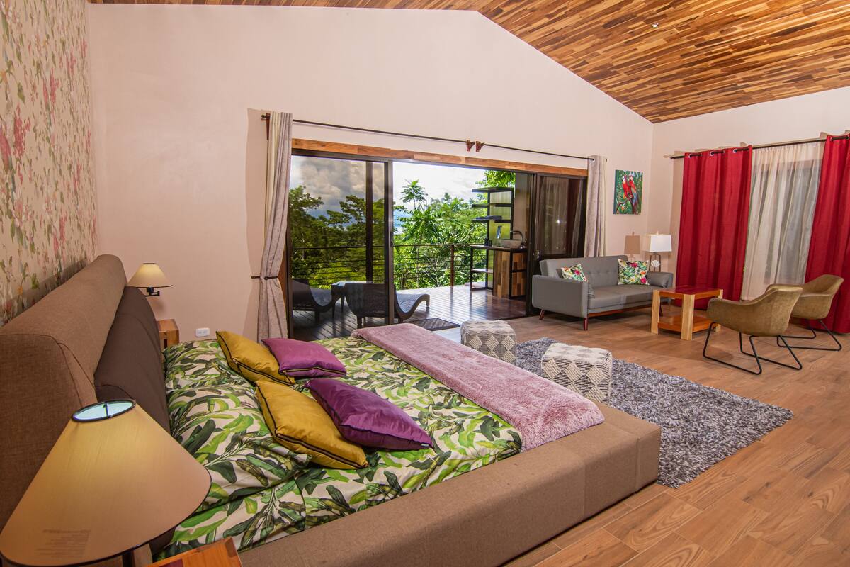 Een moderne slaapkamer met een groot bed, een bank, een klein tafeltje en een stoel. Er is een balkon met uitzicht op bomen en een rood gordijn.