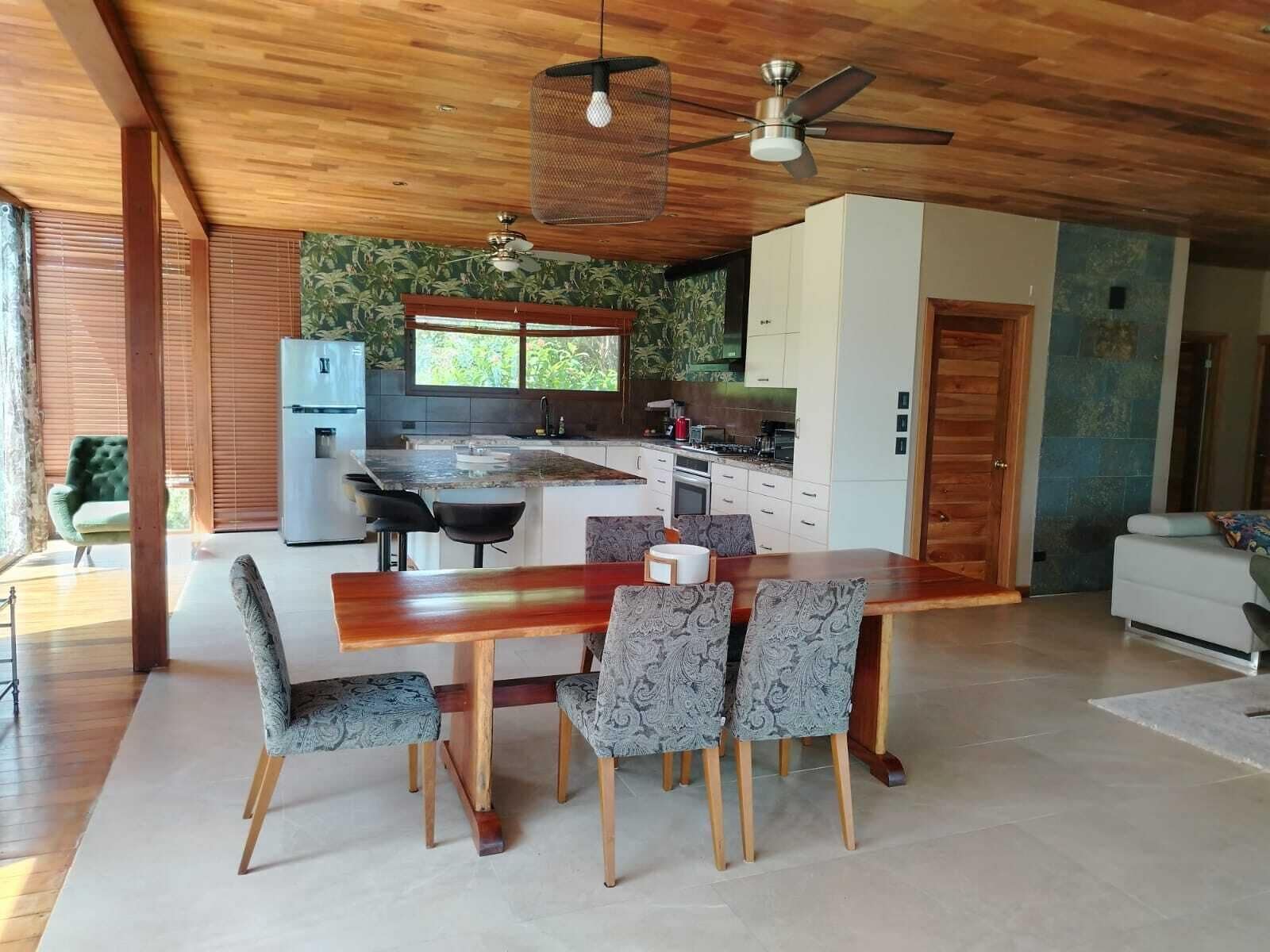 A modern kitchen with a dining area, wooden ceiling, and a large window.