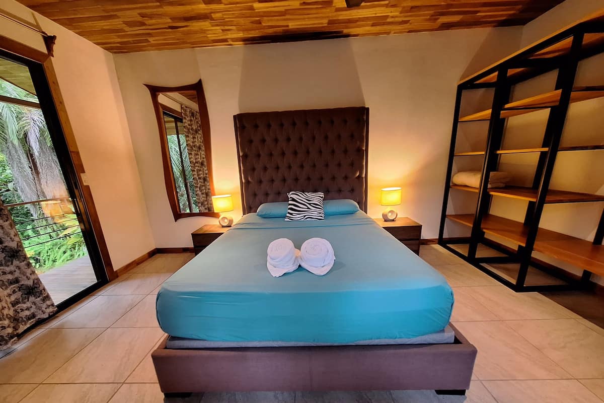 Een slaapkamer met een groot bed, twee bedlampen, een houten hoofdbord en uitzicht op groen.