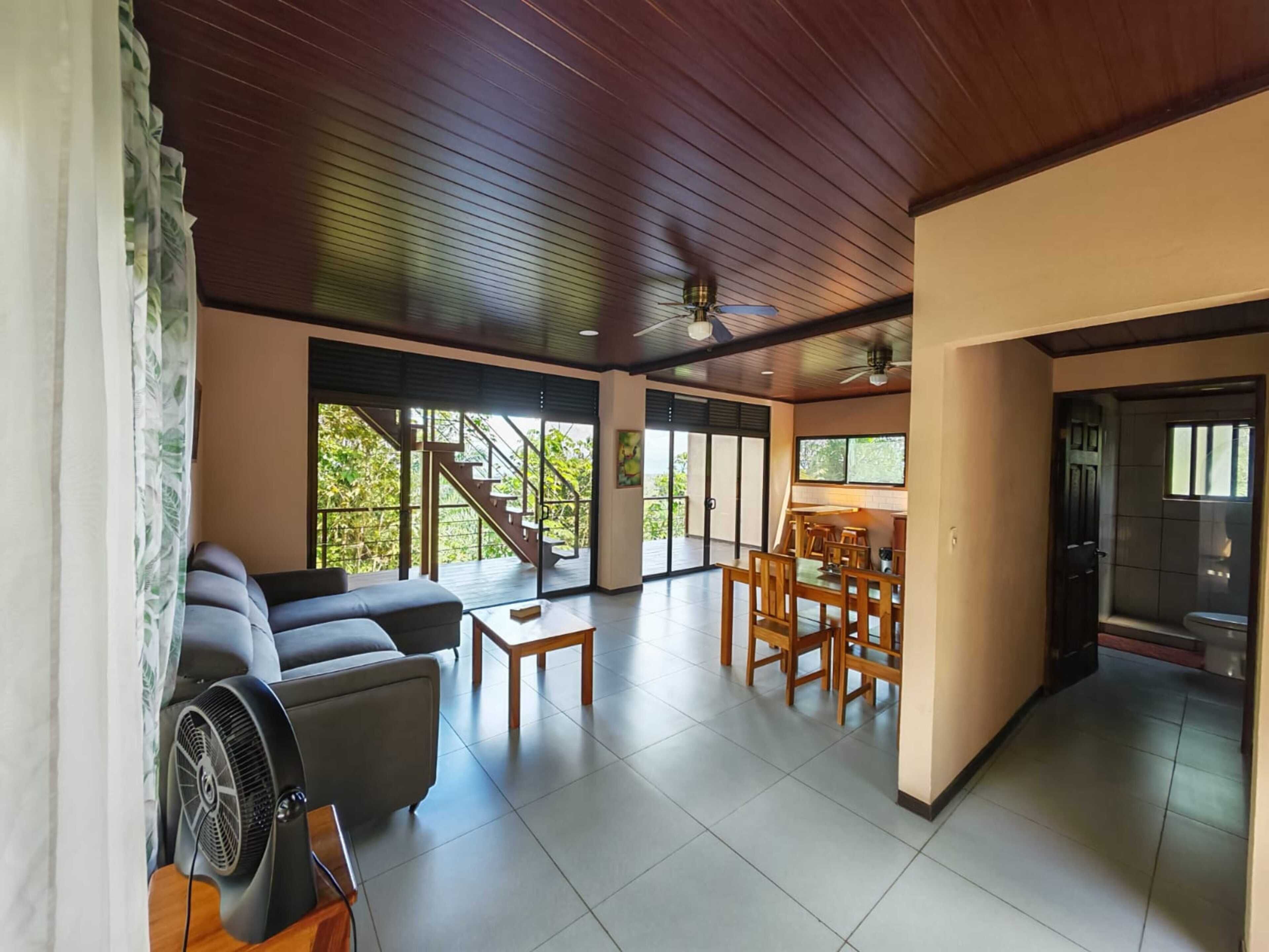 A spacious living area with a sofa, dining table, and chairs, a fan, and a staircase leading to a balcony.