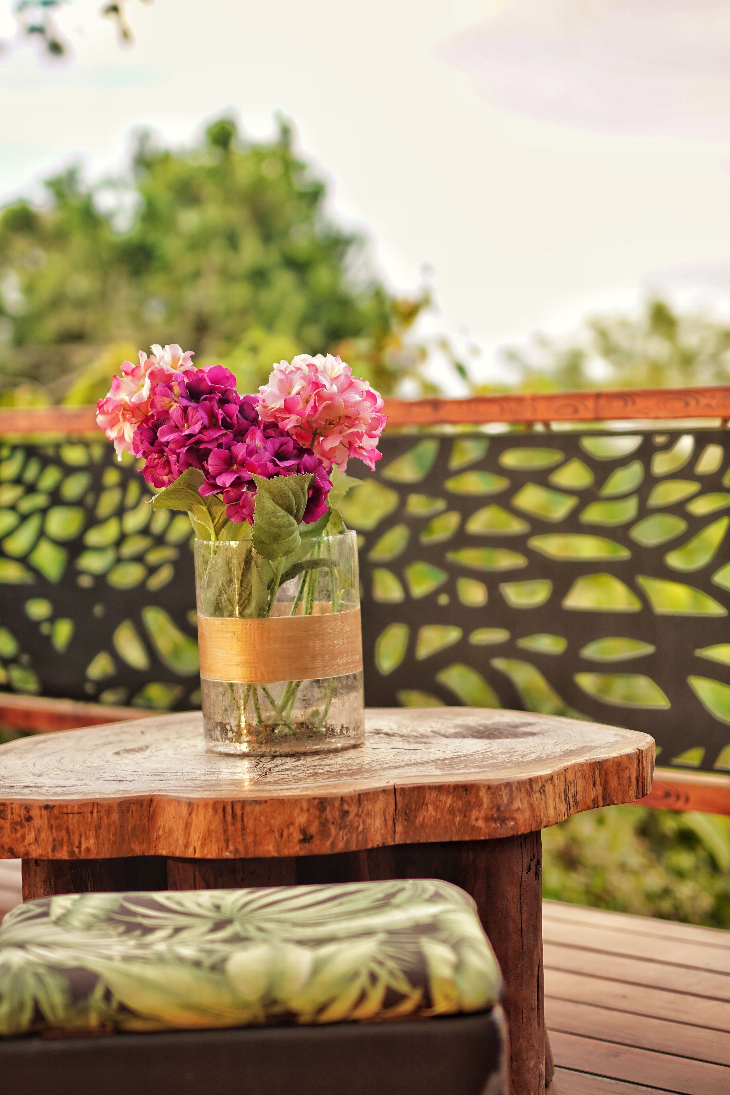A wooden table with a glass vase containing pink flowers, set on a wooden deck with a patterned cushion.