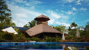 A pool area with a thatched-roof structure, lounge chairs, and umbrellas. - Corcovado Private Villas (Puerto Jiménez)