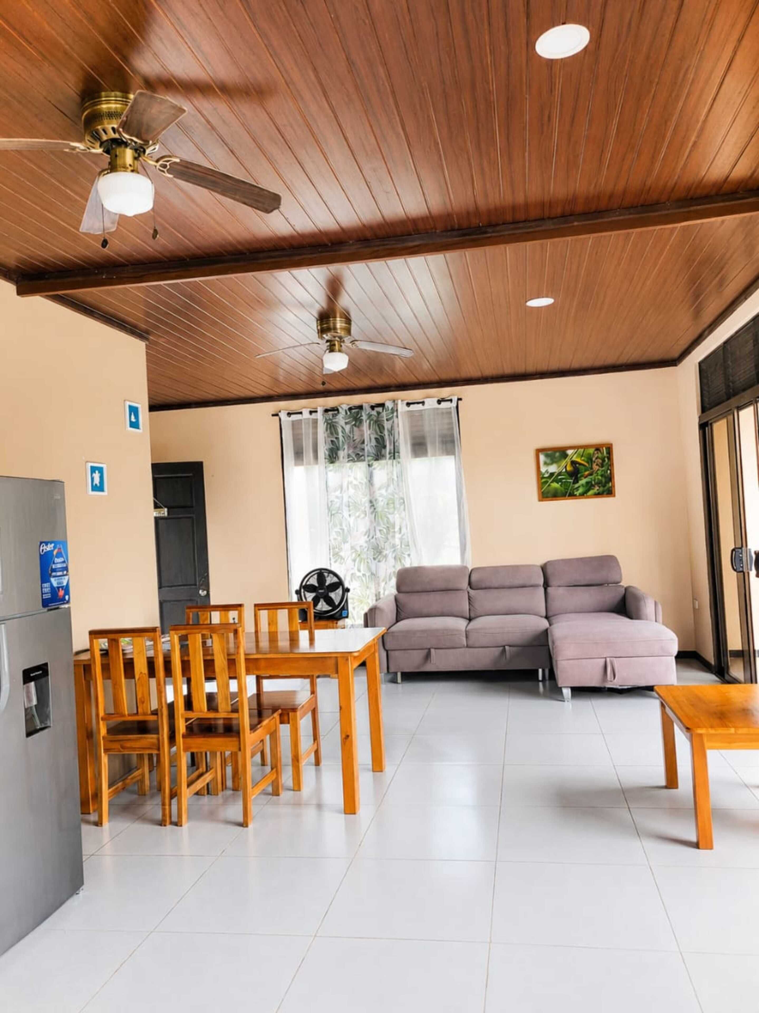 A room with a wooden ceiling, a ceiling fan, a sofa, a dining table with chairs, and a refrigerator.