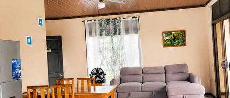 A room with a wooden ceiling, a ceiling fan, a sofa, a dining table with chairs, and a refrigerator.