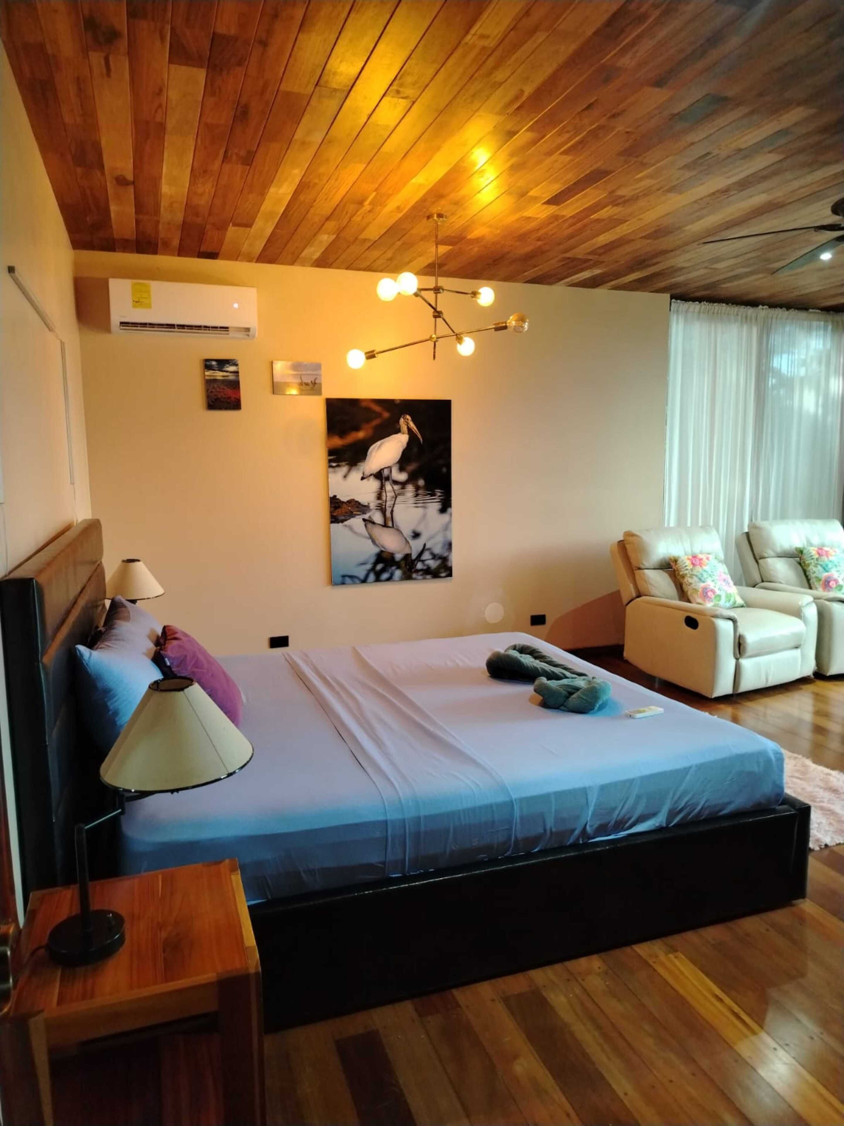 A bedroom with a wooden ceiling, a bed with a blue bedspread, a wall-mounted air conditioner, a ceiling light fixture, and a chair with floral cushions.