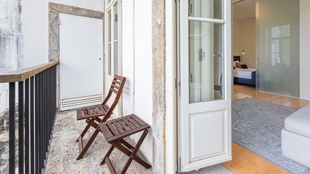 Un interior modern amb un balcó, una porta corredissa de vidre que dona a un dormitori i una petita zona de seients amb dues cadires de fusta.