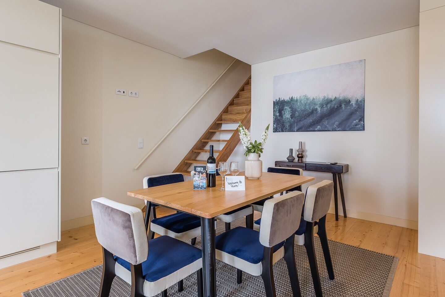 A dining area with a wooden table, blue upholstered chairs, a small potted plant, a wine bottle, and a welcome sign.