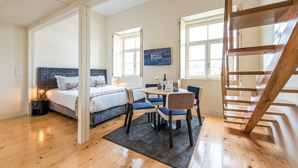 A bedroom with a bed, bedside table, lamp, and a view of the city through large windows. - GuestReady - La Maison Aires Gouveia #8 (Porto)