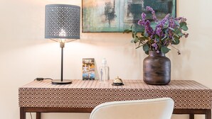 A hotel desk with a patterned tablecloth, a lamp, a vase with flowers, a framed picture, a bottle, and a small bell. - GuestReady - La Maison Aires Gouveia #8 (Porto)