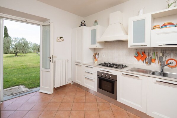 Une cuisine avec des meubles blancs, une cuisinière à gaz et une vue sur une pelouse verdoyante par une porte ouverte.
