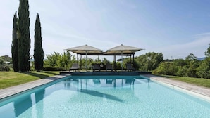 Una piscina rectangular con un deck de madera, dos altos cipreses y un pabellón con sombrillas y sillas reclinables.